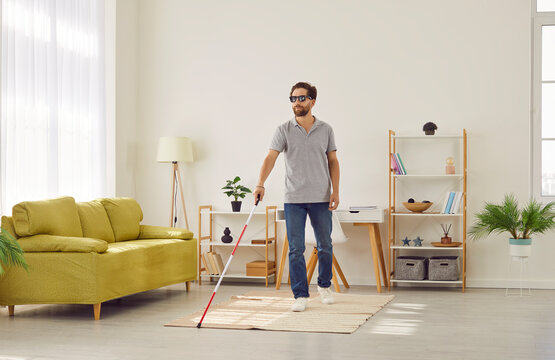 Full length portrait of blind young disabled man in casual clothes with cane walking in the living room at home. Male person with cane indoors. Blindness, disability and vision problems concept.