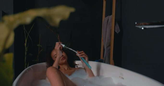 Camera reveals attractive African American female having fun blowing soap bubbles lying in a modern bathtub