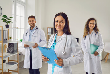 Portrait of a happy successful team of three young doctors in white uniform looking cheerful at camera and smiling while standing in hospital or clinic. Medical staff and health care concept.