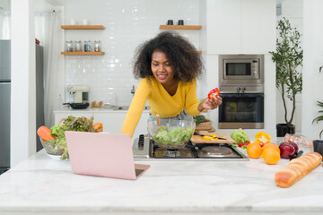 A young woman stands in a kitchen with a laptop and vegetables, likely cooking or looking up...