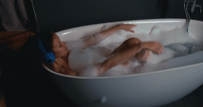 Relaxed beautiful young African American woman in wireless headphones plays with foam in stylish bath, raising hands.