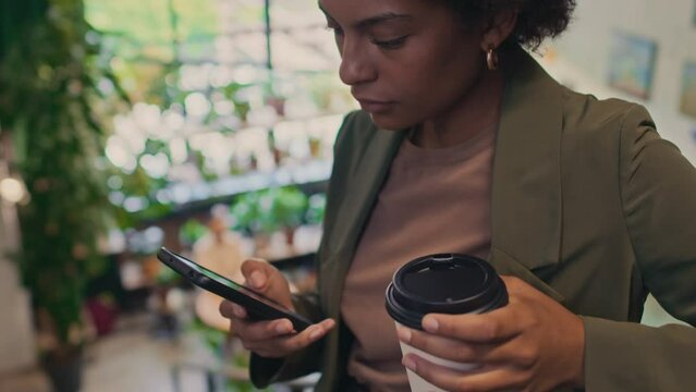 Close-up tilting shot of young African American businesswoman in trousers, blazer and t-shirt standing in office, holding takeaway coffee and typing message or email on smartphone