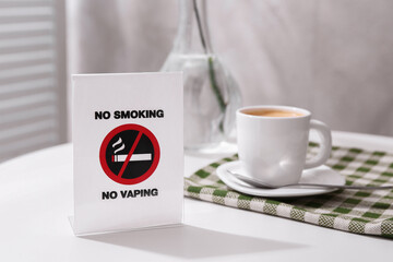 No Smoking No Vaping sign and cup of coffee on white table indoors