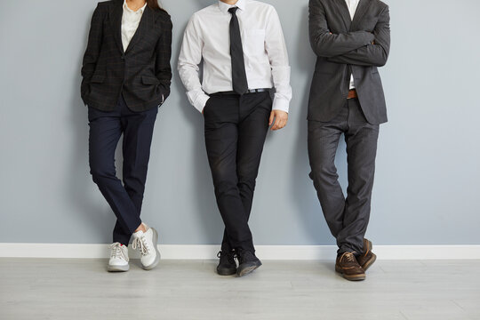 Business team in office clothes. Group of people wearing formal suits and casual shoes standing by a grey office wall and leaning on it confident, relaxed poses. Crop shot. Business, fashion concept