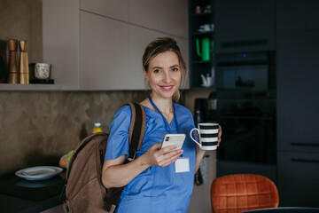 Female doctor getting ready for work, reading message on smartphone, drinking cup of coffee,...