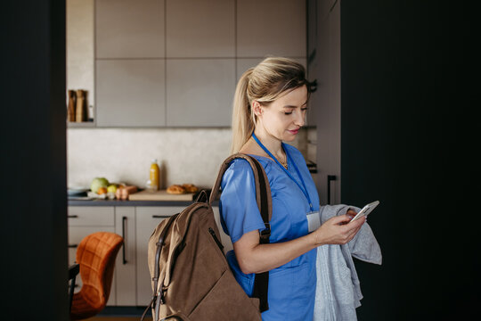 Female doctor getting ready for work, reading message on smartphone, leaving house in scrubs with backpack. Work-life balance for healthcare worker.