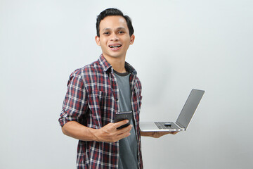 Happy asian indonesian man holding smartphone and laptop computer on isolated background