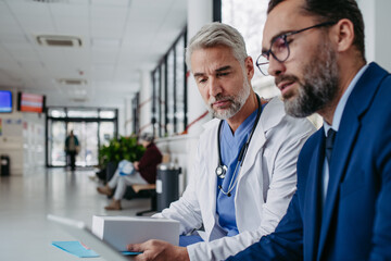 Pharmaceutical sales representative talking with doctor in medical building, presenting new medication on tablet. Hospital director, manager in modern clinic with surgeon.