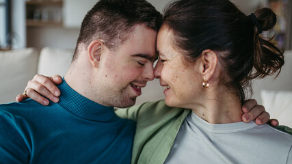 Portrait of young man with Down syndrome with his mother at home, holding, touching with foreheads....