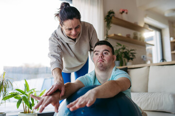 Young man with Down syndrome exercising at home with his mother on fitness ball. Workout routine for disabled man.