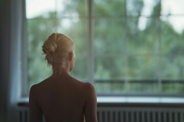 Naked young woman with blond hair looking through window