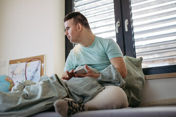 Young man with down syndrome sitting in bed, looking at smartphone in morning. Morning routine for...