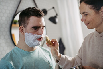 Mother applying shaving foam on son's face. Young man with down syndrome learning how to shave. © Halfpoint