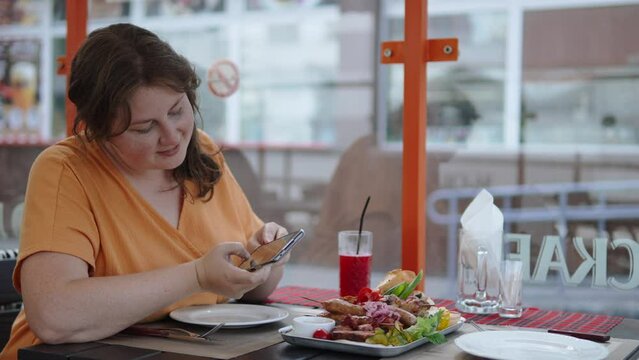 Plus size woman takes photographs of national Balkan dishes on terrace of cafe. Traveler takes pictures of appetizing assortment of grilled meats and vegetables on smartphone from different angles.