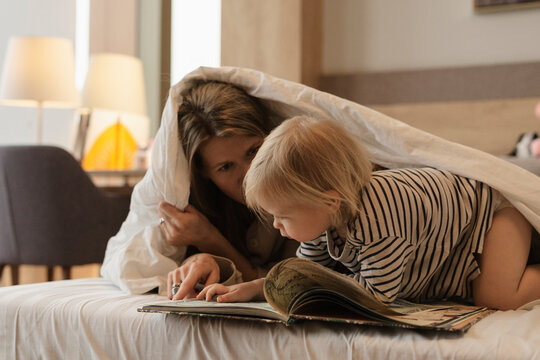 Mother Reading Book To Daughter Under Blanket In Bed At Home