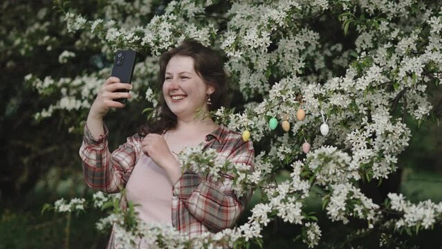 Happy woman takes selfie against backdrop of blooming spring tree. Lady with smartphone near branches with white flowers and colored decorative Easter eggs. Congratulatory video call for holiday.