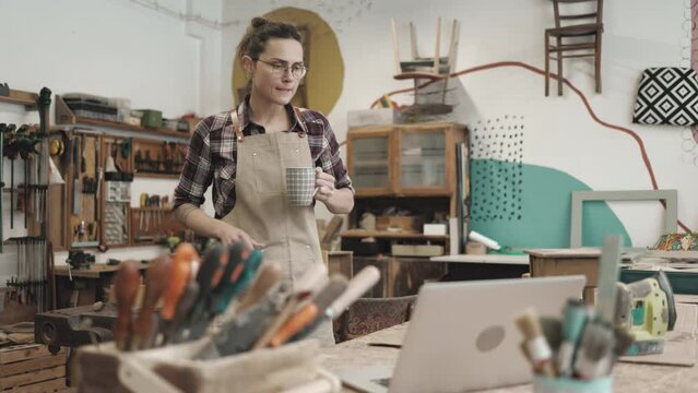 Female carpenter drinking coffee and using laptop in her workshop
