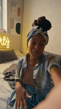 Vertical POV UGC of attractive African American lady in durag sitting in bed and talking about new apartment with neon sign Good Vibes Only on wall looking at camera and smiling