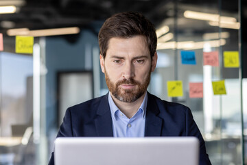 Businessman looking closely at laptop screen, mature serious man with a beard in business suit...