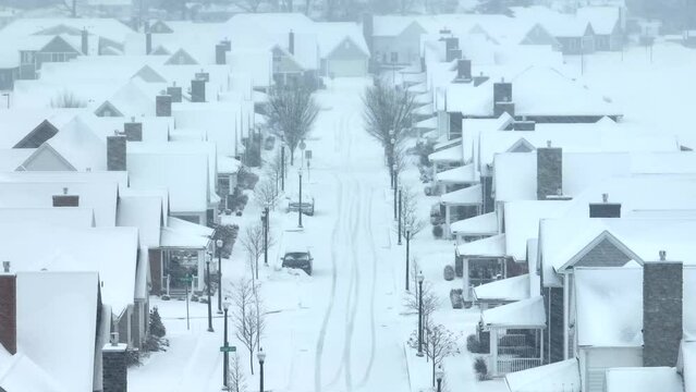 Suburban street blanketed in snow, with distinct tire tracks and houses in a row. Aerial during blizzard in USA neighborhood.