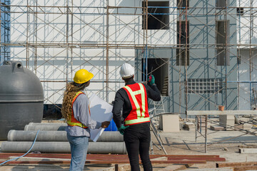 Two construction workers are standing together in front of a large building site with helmet and...
