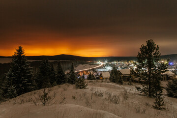 Mountain landscape with village in winter, houses and pine trees covered snow at night. Scenery of ski resort with lights.