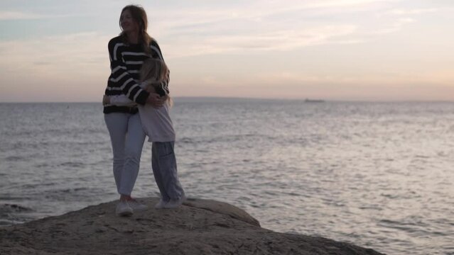 Mother and little daughter stand in an embrace by the sea in the evening