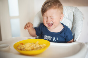 Portrait of adorable baby boy sitting on the chair and crying while eating baby food. Healthy nutrition for kids concept