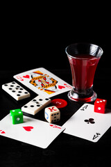 Top view of cards, dice and dominoes with glass of liquor on black background, vertical with copy space