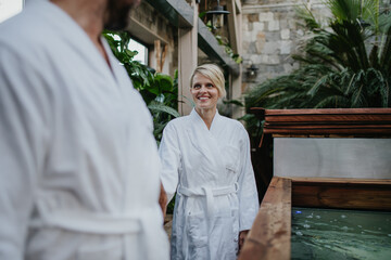 Beautiful couple in spa robes standing in hotel greenhouse, looking at each another, enjoying romantic wellness weekend in spa. Concept of Valentine's Day.