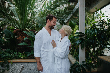 Beautiful couple in spa robes standing in hotel greenhouse, looking at each another, enjoying romantic wellness weekend in spa. Concept of Valentine's Day.