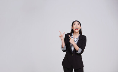 portrait of an attractive asian businesswoman in suit standing with presenting or showing hand pointing finger to copy space for product isolated on gray background.