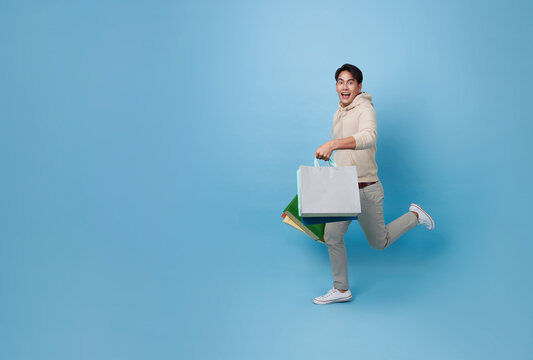 Full length young asian shopper man running and holding shopping bag feeling happy excited for discount promotion isolated on blue copy space background.