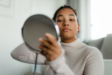 Portrait of beautiful curvy woman, looking at small round makeup mirror at home.