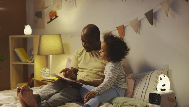 Side shot of African American dad reading and explaining story plot from cartoon book to little sleepy son in pajamas lying together in bed before sleep at night