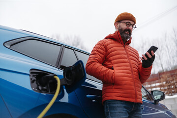 Man charging electric car during cold snowy day, using electric vehicle charging app, checking battery life, energy consumption on smart phone. Charging and driving electric vehicles during winter