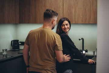 A young couple is standing in the kitchen. The guy sneaks up behind the girl and touches her waist with his hand. The girl turned to the guy and smiled - obrazy, fototapety, plakaty