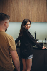 A young couple is standing in the kitchen. A guy sneaks up on a girl from behind. The girl turned to the guy.