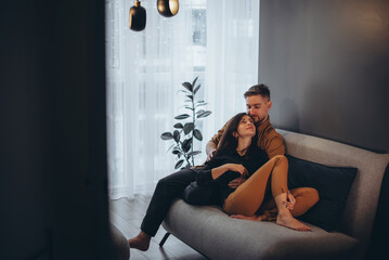 A married young couple sits on a dark sofa in a cozy apartment against the backdrop of a graphite wall and window. Couple hugging and kissing