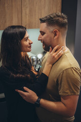 A young couple is standing in the kitchen. The girl holds the guy by the neck and looks tenderly into his eyes