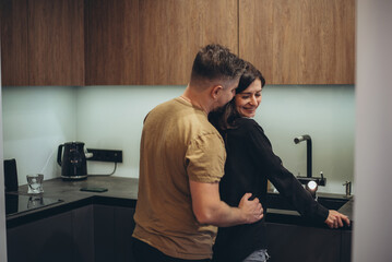 A young couple is standing in the kitchen. The guy sneaks up behind the girl and touches her waist with his hand. The girl turned to the guy and lowered her eyes in embarrassment - obrazy, fototapety, plakaty