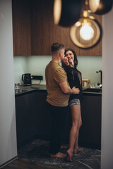 A young couple is standing in the kitchen. The guy approaches the girl from behind and touches her...