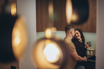 A young couple is standing in the kitchen. The guy approaches the girl from behind and touches her waist with his hand. The girl looks at the guy, smiles and holds a cup in her hands