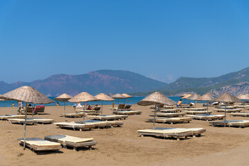 Fototapeta na wymiar Sun loungers and umbrellas on Iztuzu beach on a sunny day against the background of mountains, Turtle beach, Mediterranean Sea, Marmaris, Turkey