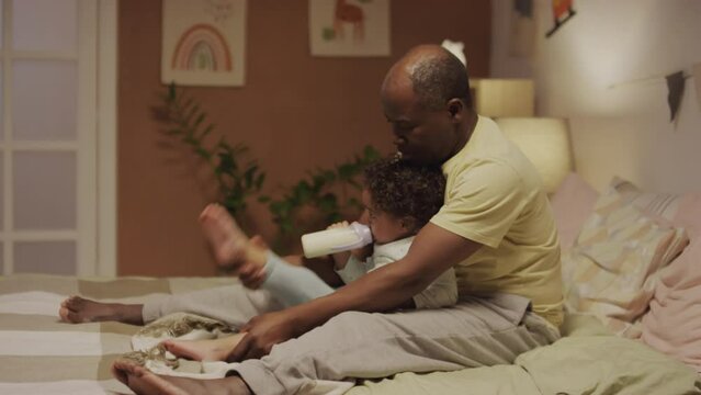 Side shot of African American dad carefully stretching Biracial baby girls legs doing exercise while daughter drinking bottle milk in cozy bedroom in evening