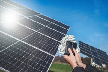 hand holding dollar bills that look out of a wallet on a background of solar panels.