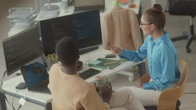 Young Black programmer drinking coffee from to go cup and listening to female colleague explaining him source code on computer monitor while sitting together at office workplace
