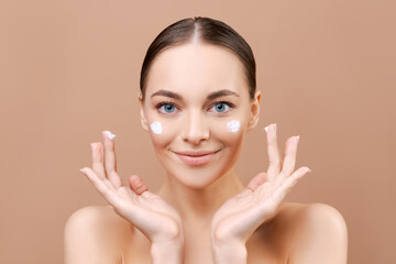 Natural beauty, young woman with cream on her face, concept facial skin care and rejuvenation on beige background in studio