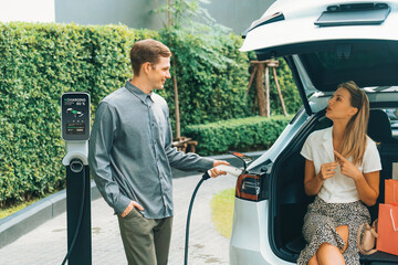 Young couple travel with EV electric car charging in green sustainable city outdoor garden in...