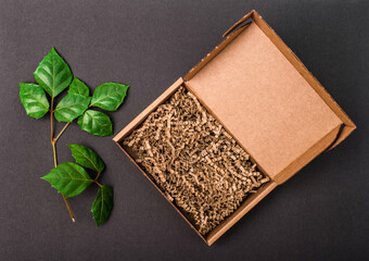 Mockup. Open cardboard box with paper filling on black background with green leaves top view.